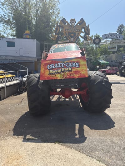 This image shows a monster truck with a sign reading "CRAZY CARS Movie Park" attached to its rear. The vehicle is parked on an asphalt surface. There are other vehicles and structures visible in the background, including what appears to be a carnival ride and a trailer. The text on the sign indicates a show time and website for the "Crazy Cars" attraction. The specific location is not explicitly stated in the image, so details about Općina Funtana, Croatia, cannot be definitively confirmed.