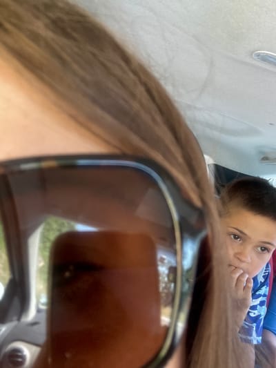 The image shows a close-up of sunglasses in the foreground, reflecting an outdoor scene. Behind the sunglasses, a young boy is visible, looking towards the camera with his hand near his mouth. He is wearing a blue and white patterned shirt. The background suggests the interior of a vehicle, possibly a car, with the ceiling and a light fixture visible. The overall scene is blurred and partially obstructed. No specific location in Lavans-lès-Saint-Claude, France can be determined from the image.