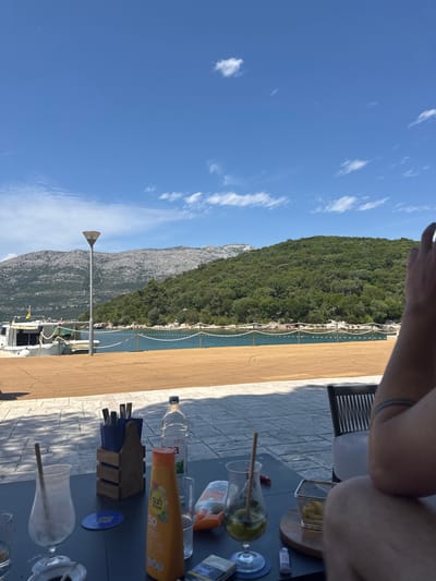 The image presents a sunny, daytime scene from an outdoor seating area, likely a cafe or restaurant, overlooking a bay in Grad Korčula, Croatia. The sky is bright blue with scattered white cumulus clouds. In the background, a rugged grey mountain range rises on the left, while a lush green, densely forested hill dominates the right. A calm, turquoise-blue body of water, possibly the Adriatic Sea, stretches across the mid-ground, separated from the foreground by a white rope barrier with buoys, within which several small figures (swimmers) are visible. A light-colored boat is docked on the left side of a wide wooden deck, partially obscured by a tall, silver conical lamp post.
In the immediate foreground, a dark-colored table is laden with various items. From left to right, there are two empty or near-empty cocktail glasses with straws, a blue coaster with a QR code, a wooden holder containing blue compartments filled with cutlery (forks, knives, spoons), and a clear plastic bottle of "Bistra" water. An orange "Sun Dance SPF 50" sunscreen bottle stands prominently, next to another smaller orange bottle. Further to the right, a glass containing a green drink with lime wedges and a straw rests on a blue coaster, beside a pack of Parliament cigarettes and a lighter. A small metal basket of french fries is placed on a round wooden board. On the far right, the tattooed right arm (featuring three dark bands) and leg of a person are visible, indicating they are seated at the table in a grey slatted chair. A shadow of another individual is cast on the light-colored, irregularly paved ground to the left of the table.