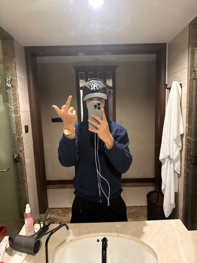 The image displays a mirror selfie taken by an individual in a bathroom setting. The person, whose upper face is obscured by a raised dark navy blue hoodie and a black New York Yankees baseball cap, holds a silver smartphone in their right hand, taking the photograph. The left hand is raised, presenting an extended middle finger. A gold-toned watch is visible on the left wrist, and a silver ring is on the ring finger of the right hand. White wired earphones are connected to the smartphone, with the wires hanging downwards. The individual is wearing black trousers.

The bathroom features light-colored marble or tiled walls and a dark wooden door frame reflected behind the person. In the foreground, a white sink with a dark faucet is set into a light-colored marble countertop. On the counter to the left of the sink, a dark gray hairdryer, a clear spray bottle containing pink liquid, and white packaged items are present. To the right, a white towel hangs on a dark wall-mounted rack, with a dark woven basket partially visible beneath it. A frosted glass shower door with a metal handle is partially visible on the far left. The specific location within Tongxiang, China, cannot be identified from the visual information provided.