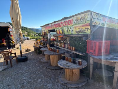 A roadside stall in the city of Ponikvica, Montenegro, displays homemade products under a clear blue sky. The stall is constructed from rustic wood and features a large banner depicting cows, sheep, and jars of produce, with the text "DOMAĆI PROIZVODI" (homemade products) prominently displayed. On wooden shelves and circular wooden spool tables, numerous glass jars filled with what appears to be honey and other liquids are arranged, alongside bottles of various sizes and colors. To the left, a person wearing a green vest and dark shorts stands near the open rear of a white van. An open umbrella stands nearby, and a plastic brown chair is placed on the gravelly ground. In the background, a white car is parked on a dirt road, and rolling green hills form the backdrop under the bright sun.