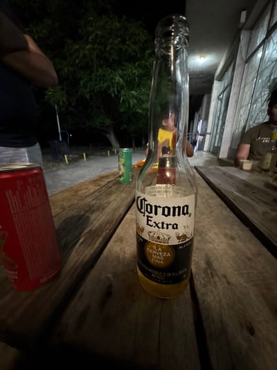 This is a low-angle, close-up shot of a bottle of Corona Extra beer on a wooden table, viewed from the side and rotated 90 degrees counter-clockwise. The bottle is mostly empty, with some liquid remaining at the bottom. The label clearly shows "Corona Extra" and "LA CERVEZA MAS FINA". In the background, out of focus, is a person wearing a brown shirt, and further back, a structure that appears to be a building with large windows or an overhang. To the left of the beer bottle, a green can is partially visible, and to the right, a red can, possibly a Coca-Cola can, is also in view. The scene appears to be outdoors, with dark green foliage visible behind the beer bottle, suggesting it might be evening or nighttime. The overall impression is a casual setting, likely a gathering or a moment of relaxation, possibly in a backyard or patio area in Gorno Aleksandrovo, Bulgaria. The lighting suggests it is either dusk or night.