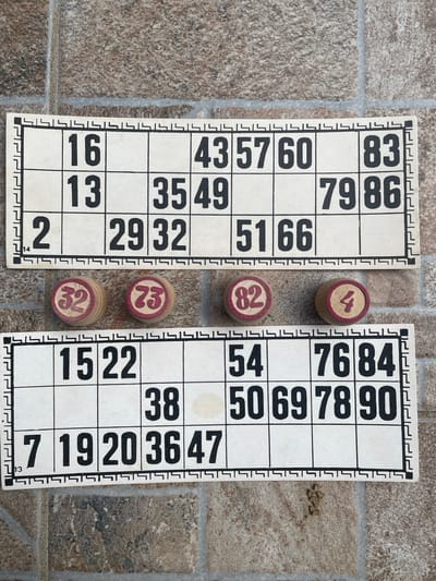 The image shows two bingo cards lying flat on a tiled surface. Between them are four bingo balls with numbers printed on them: 32, 23, 82, and 4. The bingo card on the left has the numbers 16, 13, 2, 43, 57, 35, 49, 29, 32, 60, 83, 79, 86, 51, and 66. The card on the right has the numbers 15, 22, 7, 19, 20, 36, 47, 54, 38, 50, 69, 78, 90, 76, and 84. The tiled surface has a gray and brown mottled pattern. There is no information in the image to indicate that it was taken in the city of Zhaltusha, Bulgaria.