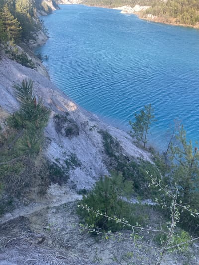 An eye-level shot shows a view from a cliff in the city of Краснасельскі сельскі Савет, Belarus. The cliff is sparsely vegetated with green foliage, with some trees growing along the edge. Below the cliff, there is a large body of water, its surface rippled with waves. The water extends to the horizon in the upper left corner of the image, giving the impression of a vast lake.