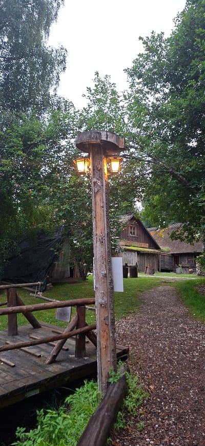 An eye-level, medium outdoor shot captures a rustic wooden lamppost with two illuminated lamps, positioned beside a wooden bridge on the left and a gravel path on the right. The lamppost, made of rough-hewn wood, has a wide, flat top and a vertical shaft with visible bark textures and a lengthwise cutout. The lamps emit a warm yellow glow.
To the left of the lamppost and bridge, lush green foliage and trees dominate the background, with a dark tarp-covered structure visible behind the bridge. The bridge itself is constructed of weathered wooden planks and railings.
To the right of the lamppost, the gravel path winds towards a collection of rustic wooden buildings. The buildings feature thatched roofs and traditional architecture, suggesting a historical or village setting. One building has a sign that appears to say "KINGSEPA KLUUB". Green grass surrounds the buildings and the path. The sky is overcast, creating a diffused light. The overall scene is evocative of a historical village or outdoor museum, likely located in Saula, Estonia, given the contextual information. There are no people or animals visible in the frame.