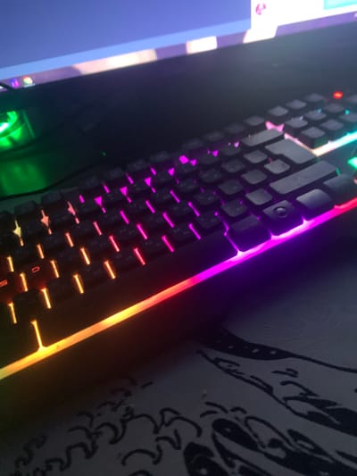 The image shows a close-up, angled view of a black gaming keyboard with RGB backlighting. The lighting transitions from green on the left side, through yellow and orange, to pink and purple on the right side. The keyboard is placed on a dark surface with a faint, abstract pattern. In the background, a portion of a computer monitor is visible, displaying a blue and purple interface, suggesting a digital environment. The overall scene depicts a gaming setup. There are no people or specific landmarks identifiable in the image that would suggest a location in Glodeni, Moldova.