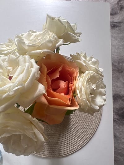 The image shows a bouquet of roses on a white surface. The roses are predominantly white, with one orange rose in the center. A round, woven mat is visible behind the roses. The background appears to be a wall with a light-colored pattern. No people are visible. There is no indication of this scene being located in Belovo, Bulgaria.