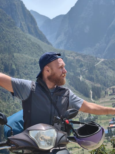 A man with a beard and wearing a baseball cap is standing next to a scooter on a mountainside in Phố Bảng Commune, Vietnam. He has his arms outstretched. He's wearing a grey t-shirt and a black vest. A blue bag is strapped to the back of the scooter, and a purple helmet is hanging on the handlebars. Mountains covered in green vegetation form the background.