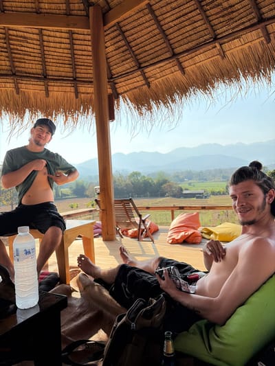 Two adult males are positioned on a wooden deck beneath a thatched roof structure in Ban Mae Ping, Thailand. The background reveals a wide vista of distant mountains and agricultural fields under a clear sky.
The male on the left, wearing a dark cap, green t-shirt, and dark shorts, is seated on a light wooden bench. He is actively lifting his t-shirt with both hands, exposing his abdomen, and pointing with his right index finger at his left nipple. He smiles directly at the camera. A clear plastic bottle labeled "Singha Water" rests on a dark table in the foreground to his left.
The male on the right, bare-chested with dark hair tied in a bun and a beard, reclines on a green beanbag cushion. He is wearing dark shorts and holds a small, rectangular object, possibly a wallet or pack of cards, in his left hand. He smiles towards the camera, with his bare feet resting on the wooden deck. A dark backpack and a brown glass bottle are visible on the deck near him.
Additional objects on the deck include an orange and a yellow beanbag cushion, and a wooden lounge chair positioned in the mid-ground. The distant landscape comprises cultivated fields and scattered trees leading up to the mountain range.