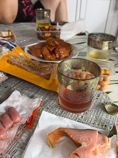 The image depicts a close-up, eye-level view of a table laden with food and drinks. The setting appears to be a casual gathering, possibly a meal or snack time. On the table, there are several items including a glass of amber-colored liquid, possibly beer or a cocktail, next to a clear plastic container filled with small, orange, spherical snacks, likely peanuts or candies. Another glass, also with amber liquid and some solid contents at the bottom, is positioned to the right. In front of the glasses, a plate holds what appears to be pieces of dried or cured meat or fish, dark reddish-brown in color. To the left, a partially opened yellow package, possibly containing crackers or breadsticks, is visible, with some of its contents peeking out. Underneath the package, a white napkin is crumpled. Further to the left, a small portion of someone's arm is visible, suggesting a person is seated at the table. In the foreground, on a white napkin, are several small, pinkish, elongated items, likely mini sausages, still in their plastic packaging. A small orange packet with some writing on it is also present near the sausages. A slice of cooked salmon with visible flakes and skin is on another white napkin to the right. A silver bottle cap lies near the glasses. The table surface is covered with a textured, greyish-green placemat. The background shows out-of-focus elements of what could be furniture or part of a room, with a white wall visible on the right. The overall impression is a spread of snacks and drinks, likely for consumption during a social occasion. The specific location in Skovorodino, Russia, cannot be determined from the visual information alone.