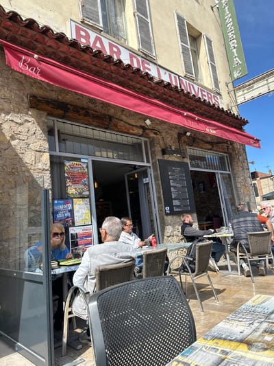 The image depicts the exterior of "Bar De L'Univers" in Mougins, France. The stone building features a red awning with "Bar" written on it. A menu board is mounted on the wall beside the entrance. Several people are seated at tables outside, appearing to be patrons of the bar. Posters and advertisements are visible in the windows. The weather appears sunny. A partial view of another building and signage is visible above.