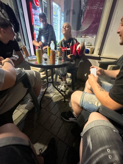 In Krivodol, Bulgaria, a group of four young adults is gathered around a circular table outside a store with illuminated refrigerators displaying beverages. The individuals appear to be engaged in a card game, with one person holding a deck of cards. Two males are seated on the left side of the frame, and a female with blonde hair, wearing a colorful jacket, is seated opposite them. A fourth male, wearing denim shorts, is seated on the right. The table is cluttered with various cans and bottles, suggesting a casual social gathering. The ground is paved with interlocking bricks, and the background reveals a store interior and a street view at night.