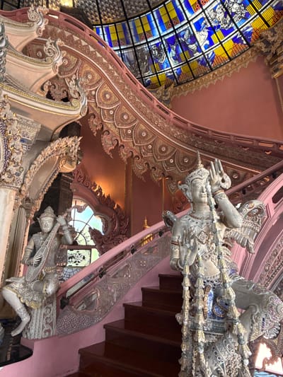 The image captures an interior architectural scene in Samrong Nuea Subdistrict Municipality, Thailand. The focal point is the ornate decoration, featuring statues of human figures with elaborate headdresses and ornamentation. These figures appear to be supporting elements of the structure. A stained-glass window in hues of blue and yellow is visible on the left side, adding color and light to the space. The interior design is characterized by intricate patterns and detail work. A pinkish-brown color palette dominates the walls and ceiling. A staircase with dark wooden steps is visible on the right side. The overall impression is one of elaborate decoration within a cultural and possibly religious context.