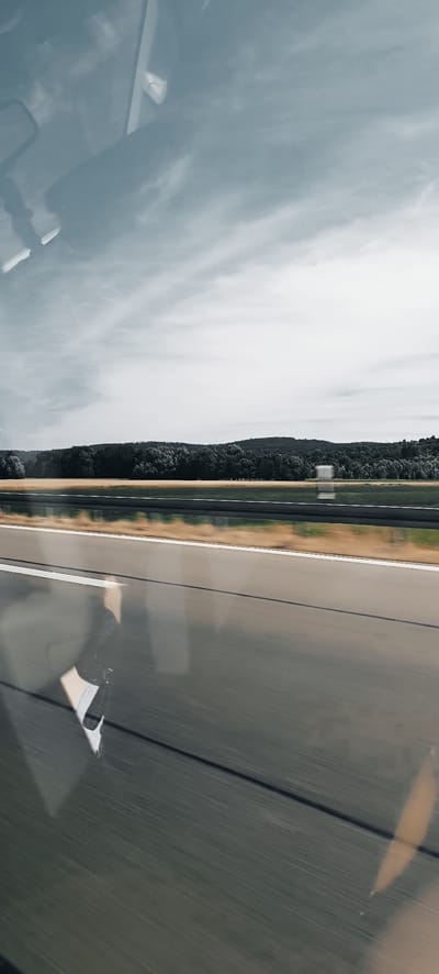 The image shows a view from a car window traveling on a highway. The foreground consists of the asphalt road with white lines, blurred due to motion. Reflections of the sky and interior of the car are visible on the road surface. A metal guardrail separates the highway from a field of yellow grass and a dark green forest in the background. The sky is overcast with wispy clouds. The scene is likely a rural or suburban area outside of a city. The provided location "Winterbach, Germany" is a town, not a city, and this image does not contain any specific landmarks to confirm this location.