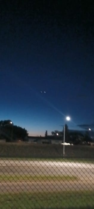 The image captures an outdoor twilight or nighttime scene within the city of Medicine Hat, Canada. The upper sky displays a dark blue hue, fading to a lighter blue with a subtle orange-pink gradient near the horizon. Two distinct bright, non-point sources of light are prominent in the upper-mid left quadrant of the sky. A faint, linear light trail is visible below these objects. In the midground, silhouetted trees and indistinct building structures are discernible against the low light of the horizon. A tall, upright street lamp with an active, bright white light fixture is positioned on the right side of the frame, casting a visible light beam diagonally towards the left. The lower third of the image is occupied by a chain-link fence, spanning across the foreground from left to right. Beyond the fence, a flat expanse of ground, appearing as a field or open area, contains a lighter-toned pathway or track. No individuals, specific actions, or interactions are depicted within the scene.