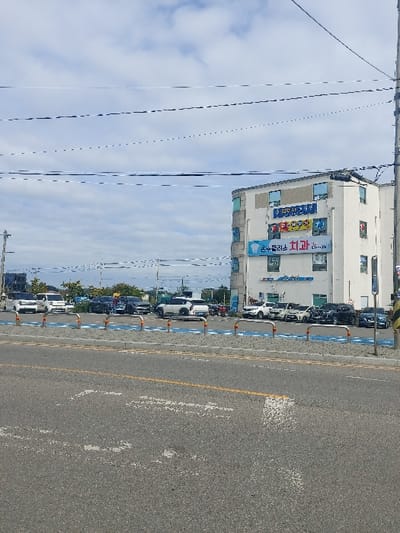 This image captures a daytime street scene in 강화군, South Korea. The foreground consists of an asphalt road displaying a faded yellow solid line and indistinct white painted road markings. Adjacent to the road, a gray curb separates it from a paved area. This paved area features blue ground markings and is delineated by approximately ten silver, short, vertical bollards.
Behind the bollards, a row of approximately ten vehicles is parked parallel to the road, facing left. These vehicles include various passenger cars and SUVs, predominantly white, black, and dark gray in color. No individuals are visible within or interacting with these vehicles.
On the right side of the frame, a multi-story building with an off-white or pale gray facade stands prominent. It features multiple windows, some with blue frames. Several large banners are affixed to its exterior. A notable blue banner on the second floor displays the Korean text "은수샘플러스 치과" (Eunsu Plus Dental Clinic). Additional signs with Korean text in various colors are present on the upper floors.
Utility poles are visible on both the left and right sides, supporting numerous overhead wires that traverse the upper portion of the frame against a partially cloudy sky. The sky is light blue with scattered white cumulus clouds. No people are visible engaged in any activity within the scene.