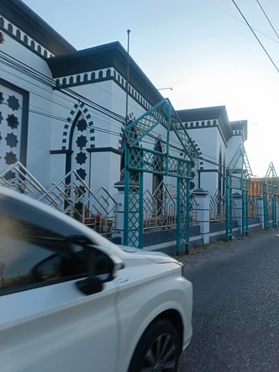 The image depicts a white car in motion, captured from a low angle on the right side of the frame, driving on a paved road. The car's front fender and a portion of its hood are visible, along with its side mirror. In the background, a white building with black architectural details and star-shaped motifs is visible. A fence with a green lattice pattern and silver accents separates the building from the road. The sky is clear and bright, suggesting daylight. The setting appears to be a street in Luwoo, Indonesia, characterized by its tropical sunlight and the style of the building. The scene is static, with the only implied action being the car's movement.