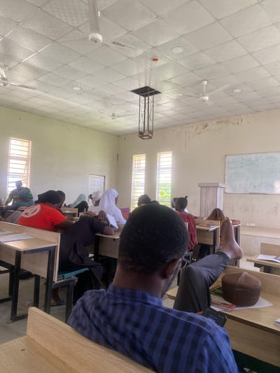 The image shows a group of people sitting in what appears to be a classroom or lecture hall in Yali, Nigeria. The room has a tiled ceiling with several ceiling fans, and windows that let in natural light, suggesting it is daytime. The walls are a light color, and there's a whiteboard in the background with some writing on it.
The subjects are primarily students seated at desks, facing towards the front of the room. Some are looking down, possibly at notes or a device, while others are looking forward. The activity taking place is likely a lecture or class session, given the setting and the attentive postures of some individuals.
Notable details include a person in the foreground wearing a blue and black striped shirt, with their back to the camera. To their left, another person is wearing a brown kufi. In the middle ground, several individuals are visible, including one wearing a white hijab and another in an orange t-shirt. There are desks made of wood and metal. The overall impression is one of a learning environment. The lighting suggests it is daytime, and the weather is not discernible from the image. The emotion conveyed is one of concentration and engagement in the educational activity.
There is some faint writing on the whiteboard in the background, but it is too blurry to be legible. A phone screen is visible in the foreground, displaying the word "ZAKIYAA."