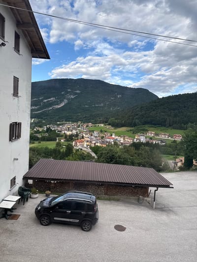 This is a photograph taken from an elevated perspective, looking down onto a small town nestled in a valley at the base of a lush, tree-covered mountain. The sky is partly cloudy, with patches of blue visible between white, fluffy clouds. The sun appears to be shining, suggesting it is daytime.
In the foreground, the edge of a white building with wooden window shutters is visible. To the right of this building, a dark-colored car is parked on an asphalt surface. Adjacent to the car is a covered structure with a brown, corrugated roof, and stacks of firewood underneath. Beyond the car and the covered structure, the town unfolds in the valley. The buildings are a mix of colors, including white, yellow, and red, and they are clustered along winding roads. The town is surrounded by green fields and trees.
The mountain that dominates the background is densely forested, with some patches of rock visible on its slopes. The overall scene depicts a tranquil, rural setting in a mountainous region. The time of day appears to be afternoon, given the angle of the sunlight. There is no visible text in the image. The location context provided is Cavedago, Italy, which aligns with the European architectural style of the buildings and the mountainous landscape.