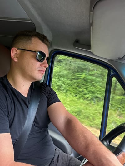 A man with sunglasses on is seen sleeping in a vehicle. He is wearing a black t-shirt and is buckled with a seatbelt. The background seen through the window is out of focus, showing green foliage, suggesting an outdoor setting, possibly a rural area. The image appears to be taken during the day, with natural light illuminating the scene. The man's face is relaxed, indicating he is asleep or resting. The interior of the vehicle, including a sun visor and part of the steering wheel, are visible, suggesting the man is a passenger or driver. The crops provide close-ups of the man's head, arm, and clothing, as well as the interior of the vehicle and the view outside. No visible text is present in the image.