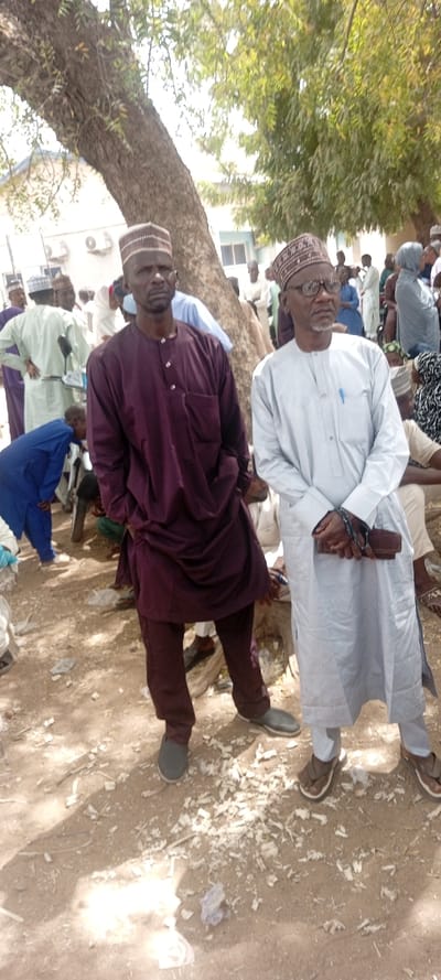 The image depicts an outdoor gathering in Sani Mai Nagge, Nigeria, under the shade of a large, mature tree with extensive green foliage. In the foreground, two men stand facing the viewer. The man on the left, of dark complexion, wears a dark purple long-sleeved tunic and matching trousers, topped with a patterned brown and white kufi cap; his hands are placed in his pockets. The man on the right, of lighter complexion, wears glasses, a light grey long-sleeved tunic with a blue pen in the left chest pocket, and a patterned brown and white kufi cap; he holds a brown rectangular object, possibly a wallet or pouch, in his left hand, and prayer beads are visible on his left wrist.
Behind these two individuals, a crowd of numerous people is visible, predominantly men in various traditional Nigerian garments and caps, alongside some women in head coverings. They appear to be standing and conversing in small groups. The ground beneath the gathering is unpaved dirt, uneven, and covered with scattered white fibrous debris and general litter. In the background, behind the crowd and partially obscured by the tree, a light-colored building with multiple windows and external air conditioning units mounted on its facade is discernible. The scene is illuminated by sunlight, casting shadows on the ground.