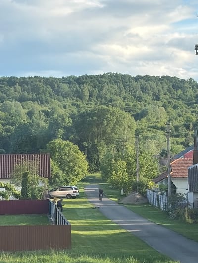 The image depicts a sunlit, rural scene in Hanniv, Ukraine. A paved road, bordered by grassy areas and fences, runs through the center of the frame, leading towards a verdant, tree-covered hill in the background. On the left side of the road, a light-colored minivan is parked near a building with a red corrugated roof. Further down the road, a person is cycling, and another person is standing near the fence on the left side of the road. On the right side of the road, houses with red-tiled roofs and white walls are visible, separated by a fence. The sky above is a mix of blue and white clouds.