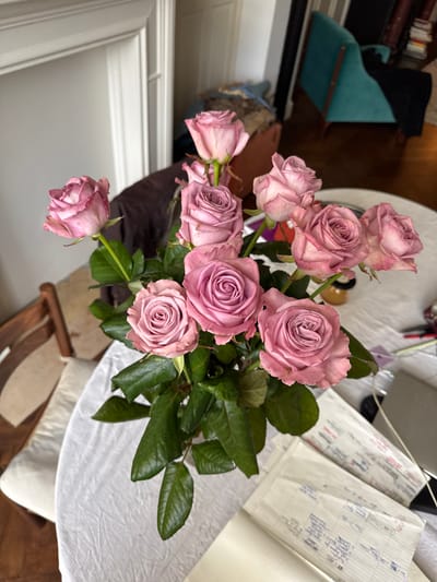 This image displays an indoor setting within a residence in Saint-Gilles - Sint-Gillis, Belgium. The central subject is a bouquet of approximately twelve light purple-pink roses with prominent green stems and leaves, contained within an unidentifiable vase on a white, textured tablecloth. The table, likely round, features a small, amber-colored jar behind the floral arrangement and a stack of printed white papers alongside a dark, rectangular object to the right foreground. In the background to the left, a wooden chair with a light-colored cushion is partially visible, adjacent to a white wall section featuring architectural molding. The background to the right reveals a teal upholstered armchair with visible wooden framing situated on a dark wooden floor. A bookshelf containing various books is dimly visible beyond the armchair. No individuals or specific actions are depicted.
