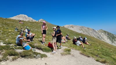 The image depicts a group of people resting on a grassy mountain slope under a clear blue sky. The terrain is uneven and rocky, with sparse vegetation. In the background, rugged mountains stretch across the horizon. The people are engaged in various activities such as sitting, standing, and conversing. Some are wearing casual clothing, while others appear to be dressed for hiking. Several individuals are wearing hats, and some have backpacks. The overall atmosphere suggests a leisurely break during an outdoor excursion. The specific location in L'Aquila, Italy cannot be determined from the image alone, as mountain landscapes are a common feature of the region.