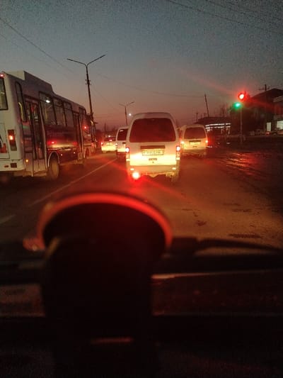 A street scene in Charkhin, Uzbekistan, is depicted at twilight or early night, viewed from the interior of a vehicle. The foreground is partially obscured by a dark, semicircular object, likely a dashboard component, emitting a red glow. An urban road occupies the midground, featuring vehicular traffic. On the left, a white passenger bus displays "Pulkerafon" on its side with interior lights illuminated. In the center and right, a queue of white vans or minivans is stationary or moving slowly, their rear lights illuminated. The lead van displays Uzbek license plate "M 931 HA" and has illuminated red taillights and white reverse lights. The road surface is wet, reflecting vehicle and ambient lights. On the right, a traffic light shows a prominent green signal and a fainter red signal above it. The background includes indistinct buildings with illuminated elements, utility poles, power lines, and sparse, bare trees against a dark blue-grey sky. No individuals are clearly discernible.