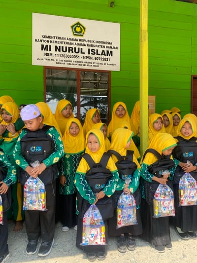In Mekar Raya, Indonesia, a group of children in matching green and yellow Islamic school uniforms stand in front of a vibrant green building. The building features a white sign with black text, identifying the institution as "KEMENTERIAN AGAMA REPUBLIK INDONESIA KANTOR KEMENTERIAN AGAMA KABUPATEN BANJAR MI NURUL ISLAM". Below this, an address is listed: "JL. A. YANI KM. 11.200 HANDIL II BANYU HIRANG RT. 02 NO. 67 GAMBUT BANJAR - KALIMANTAN SELATAN 70652".
The children, all wearing yellow hijabs, are holding transparent plastic bags filled with school supplies. One boy in the foreground, wearing a white and purple patterned cap, clutches a bag with both hands. The other children also hold their bags, some looking towards the camera, others looking away. Behind them, wooden window frames are visible, suggesting the entrance to the school. The overall scene depicts a charitable distribution or a school event.