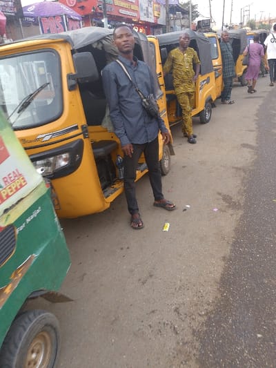 A man stands next to a yellow three-wheeled taxi. He wears a dark blue shirt, dark trousers, and sandals. A black bag hangs across his body. Other taxis and people are in the background. The scene appears to be a busy street in a city. A green taxi is partially visible on the left side of the frame.