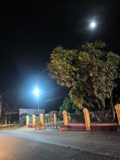 This nocturnal image, set in Meuse, Indonesia, portrays a quiet street scene illuminated by both natural moonlight and artificial street lighting. The sky is a deep, dark black, with a bright, full moon prominently positioned in the upper right, casting a soft glow through thin cloud cover. Several faint stars are discernible in the vast expanse of the night sky.
A large, dense-canopied tree, possibly a mango tree, dominates the right side of the frame, its textured leaves and branches illuminated by ambient light. Running across the upper and middle sections of the image are multiple utility wires, faintly visible against the dark sky.
On the left, a powerful streetlamp emits a brilliant, blue-white light, creating a significant lens flare and acting as a primary light source for the foreground and mid-ground. Below this light source is a segment of a paved road or street, visible in the foreground, showing reflective textures from the scattered light.
Further back, an ornate entrance to a property is visible, featuring a decorative metal gate flanked by substantial pillars with yellowish-orange tops and reddish-brown brick bases. Small plants in pots or flowerbeds are placed near the gate's pillars. Beyond the gate, a dimly lit building structure and additional foliage can be glimpsed in the background.
The scene is devoid of people or any discernible activity, conveying a sense of stillness and calm. No visible text is present in the image.