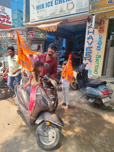 This image captures a bright, sunlit street scene in Anantapur, India, identifiable by the "AP 39C" vehicle registration plate. In the foreground, a young man wearing glasses and a maroon collared shirt smiles directly at the camera. He stands behind a young girl seated on a brown Honda scooter, which has an orange flag bearing "जय श्री राम" (Jai Shri Ram) attached to its front and another held by the girl. The girl wears a cream top and a pink skirt, looking towards the right. To the girl's right, a young boy in a white shirt and light grey pants stands holding another identical orange "Jai Shri Ram" flag. On the far left, an adult male in a light green shirt and blue jeans stands near a red motorcycle, looking off-camera. The street surface is unpaved, dusty, and contains scattered debris. In the background, several shops line the street, including a "MIX ICE CREAM" establishment advertising "Faluda," "Kulfi," and "ORDER TAKEN FOR MARRIAGE AND PARTY." Another prominent vertical sign reads "MAHAVEER" in blue lettering. A grey scooter is parked on the right side of the frame, displaying the license plate "AP 39C 84360." Several other individuals are visible in the background, some inside the shops.