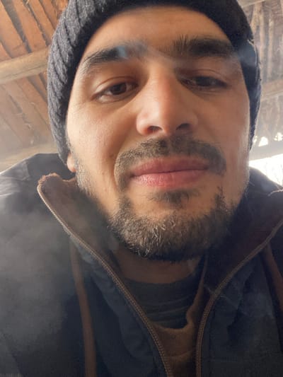 A low-angle, close-up selfie of a person with light skin, dark hair, and a stubbly beard. The individual wears a dark beanie and a dark jacket with a brown inner lining near the zipper. The person's expression is neutral, and they appear to be looking at the camera. Behind them is a blurred background of what looks like wooden beams, possibly a ceiling or structure. The lighting is soft and diffused, giving the image an overall natural and somewhat hazy appearance.