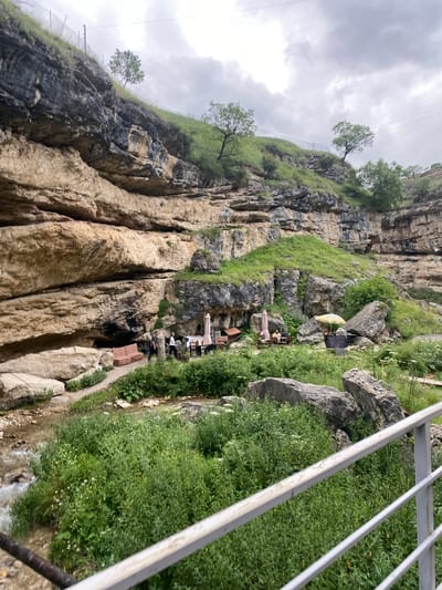 The image depicts a natural landscape with a cliff face, vegetation, and a stream, likely located in the city of Salta, Russia. A path winds along the base of the cliff, leading to an area with outdoor seating, including tables and umbrellas, suggesting a cafe or restaurant. Several people are visible in this seating area. The stream flows through lush green vegetation in the foreground, with large rocks scattered throughout. The cliff itself is layered with varying shades of rock and patches of greenery. The sky is overcast. A metal railing is present in the lower right corner of the image, suggesting a viewing platform or walkway.