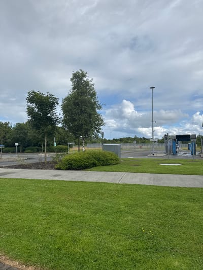 The image depicts an outdoor scene with a clear sky and fluffy white clouds. On the right side of the frame, a neatly mowed green lawn runs alongside a paved walkway. Further in the background, a car wash facility is visible with its blue brushes and structure. Trees are scattered throughout the scene, with some larger trees on the left and a dense green hedge on the right. A tall pole with a light fixture stands prominently in the middle left of the image, with security cameras mounted on it. Several signs are visible, including one that reads "CASH ONLY" and another with a symbol of a person walking. The overall setting appears to be a public space or a commercial area, possibly a car wash or a service station. The time of day appears to be daytime, given the bright lighting. The weather is partly cloudy, indicating a pleasant day.