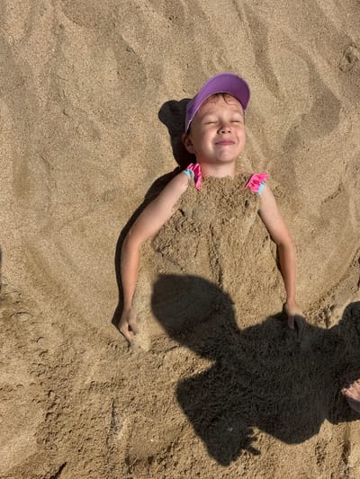A young girl with a purple hat and pink swimsuit is buried in sand, with only her head and arms visible. She is lying on her back with her eyes closed and a smile on her face, appearing happy and relaxed. Her arms are outstretched, and her hands are resting on the sand. The sand is a light brown color and has been sculpted around her body. There is a large shadow cast on the sand to the right of the girl, suggesting it is a sunny day. The background is also sand, with some footprints visible. This is a candid shot of a child enjoying a day at the beach. The location is Blagoveschenskaya, Russia.