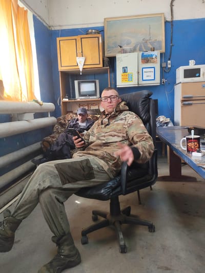 In a dimly lit room, likely a break room or office, two men are present. One man, wearing glasses and a camouflage jacket, sits in a black leather office chair, holding a mobile phone in his right hand and looking towards the camera. His left hand is raised, possibly in a gesture. The other man is seated on a patterned sofa, wearing a cap and a dark camouflage jacket, and appears to be looking at his phone as well.

The room's walls are painted a striking blue. A window with orange curtains is visible on the left. In the background, a wooden cabinet is mounted on the wall, with a small pennant hanging from it. Below the cabinet, a vintage television set is placed on a shelf. Further to the right, a microwave oven sits atop another unit, with a blue and white electrical panel box mounted on the wall between them. A framed painting hangs above the electrical panel. A mug with a spoon is on a table in the foreground to the right.

The overall impression is one of a utilitarian space, possibly in a remote or industrial setting. Based on the visual cues of the painting and the calendar visible in the image, which indicates "2019", and the context of the village of Termal'nyy, Russia, this scene could depict a break area within a facility in that location. The environment suggests a practical, no-frills setup.