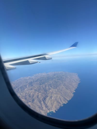 From the window of an aircraft, the wing is visible against a clear blue sky with a view of land that appears to be the city of Paiania Municipal Unit, Greece. The landscape is uneven, consisting of cliffs, rocky and elevated terrain, and a small amount of vegetation. There is also a body of water that borders the land.