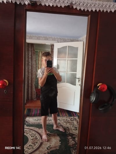 This is a full-length mirror selfie taken by a young boy in what appears to be a bedroom. He is standing in front of a closet with a dark wood finish, to his left is a closet door with a decorative red handle and keyhole. To his right, headphones are hanging on the closet door. He is wearing a white t-shirt and black shorts. The boy is holding a smartphone and taking a picture of himself. In the background, a white door with glass panes is visible, along with a patterned rug on the floor and curtains on a window. The time stamp in the bottom right corner indicates the image was taken on July 1, 2026, at 12:46 PM. The text "REDMI 14C" is visible in the bottom left corner, which is likely the model of the phone used to take the photo. The setting seems to be indoors, possibly a home in Pepeni, Moldova, given the location context. The lighting suggests it is daytime, and the overall mood is casual and personal.