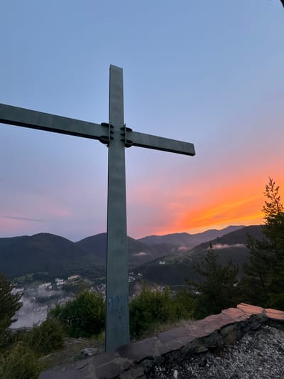 A dark, angular cross stands prominently in the foreground, its metallic structure stark against a gradient sky. The sky transitions from a soft, pale blue on the left to vibrant hues of orange and pink near the horizon, suggesting either sunrise or sunset. Behind the cross, rolling hills and mountains are shrouded in a dusky light, with some appearing to be covered in a light mist. Scattered lights from a village or town can be seen nestled in the valley below, adding a sense of human presence to the natural landscape. The terrain on the right of the image is rough and rocky, with some greenery encroaching, hinting at a mountainous or elevated viewpoint. The overall mood is serene and contemplative, amplified by the dramatic sky. The time of day is likely dawn or dusk. There is some visible graffiti on the horizontal beam of the cross, appearing as blue markings.