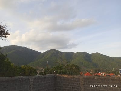 The scene, located in Namanga, Kenya, was recorded at 17:55 on November 21, 2025. The foreground is occupied by a gray concrete block wall, exhibiting an unfinished or utilitarian appearance. Immediately behind this wall, a security fence topped with razor wire extends across the mid-ground. Further back, a town or settlement is visible, characterized by buildings with red-tiled roofs amidst green foliage. A prominent red and white striped pole, possibly a telecommunications mast, stands vertically in the mid-ground. A small solar panel array is mounted on the right, above the concrete wall. The background consists of multiple densely vegetated hills, forming a continuous ridge. The sky above is partly cloudy, displaying patches of pale blue and scattered gray clouds. A barren tree branch partially enters the frame from the upper left. No people are present or engaged in any activity.