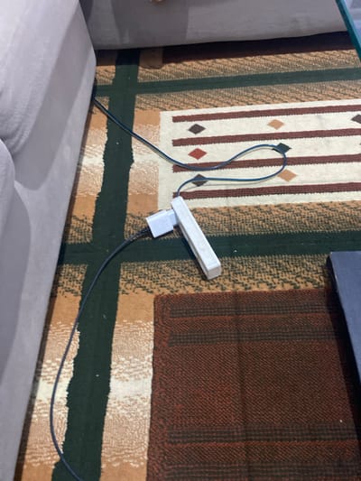 The image shows a close-up view of a rug with a geometric pattern of vertical stripes in various colors, including white, brown, dark green, and a reddish-brown. A white power strip with multiple outlets is plugged into a white adapter, which is connected to a black cable. A thin blue cable is also visible, looped over the power strip. The rug appears to be on the floor, next to a light gray sofa or couch. Part of a dark blue object, possibly another piece of furniture, is visible at the top of the image. The scene is indoors, with artificial lighting. There is no visible text in the image. The focus is on the power strip and cables resting on the patterned rug. The overall impression is one of a domestic setting, possibly a living room.