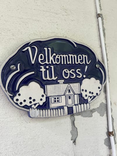 A decorative ceramic plaque hangs on a textured white wall, next to a white pipe. The plaque is dark blue with white lettering and a white illustration of a house with a fence, trees, and what appears to be a rainbow. The text on the plaque reads "Velkommen til oss!". The plaque is secured to the wall with a screw visible on the left side. The wall itself is weathered, with some peeling paint or plaster. The overall scene is an exterior view of a building's facade in Kongsberg, Norway.