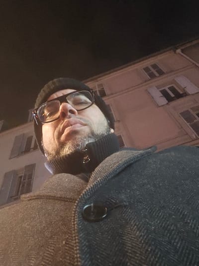 The image captures an adult male subject outdoors at night in Le Kremlin-Bicêtre, France. The individual, viewed from a low-angle perspective, is looking directly at the camera. He is wearing a black knit beanie, dark-rimmed eyeglasses, a black ribbed turtleneck, and a dark grey herringbone patterned overcoat with a single visible black button. He has a short, scruffy beard and mustache. In the background, a multi-story building with light-colored walls and multiple windows featuring closed louvered shutters is dimly visible, out of focus, under artificial illumination. The sky above is dark, indicative of nighttime.