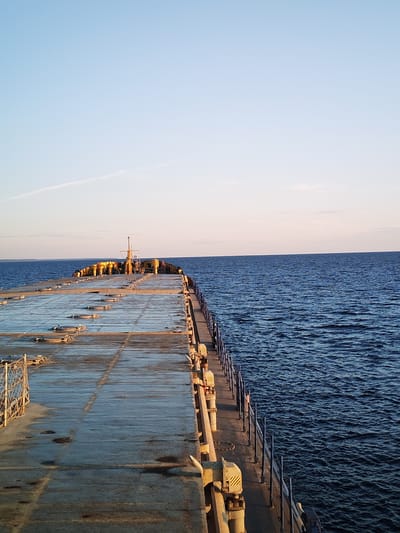 The image depicts a view from the deck of a vessel, looking towards the stern. The deck is wet and appears to be made of concrete or a similar material, with numerous circular hatches visible. A metal railing runs along the starboard side of the deck. In the distance, the stern of the vessel is visible with yellow structures and a mast. The vessel is on a large body of water, likely the sea, with a clear sky overhead. The horizon is visible in the distance, with a faint cloud or contrail in the sky. The scene is devoid of any people or specific landmarks to indicate a precise location in Kakkarovo, Russia. The lighting suggests it is either early morning or late afternoon.