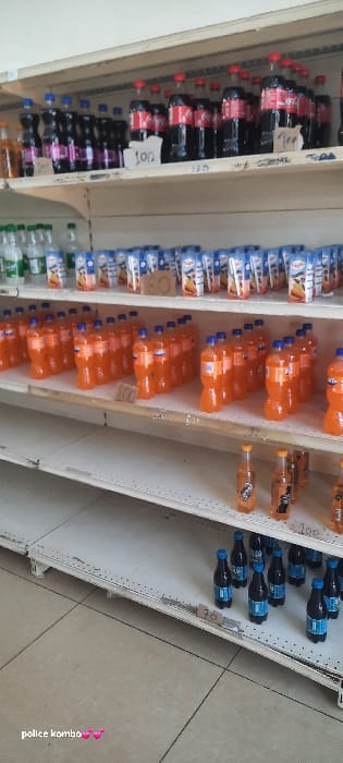 A tilted shot reveals store shelves stocked with a variety of bottled beverages. Coca-Cola bottles occupy the top left, followed by blue-and-white labeled drinks. Orange-colored soda fills the middle shelves. Smaller bottles of orange soda are beside them. The bottom shelves display water bottles and an additional assortment of blue-capped drinks. Price tags are visible on some shelves, providing a glimpse into the cost of each item.
