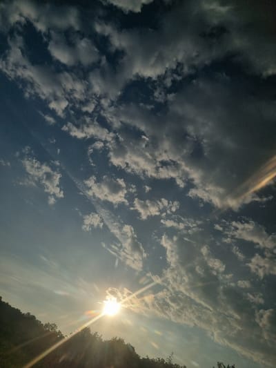 The image depicts a view of the sky with the sun setting or rising over a silhouetted tree line. The sky is a mix of blue and dark clouds, with the sun casting bright rays and lens flares across the scene. A faint rainbow-like atmospheric phenomenon is visible on the right side of the image. The perspective is angled, looking upwards towards the sky from a lower vantage point, suggesting a landscape or outdoor setting. The location is Makresh, Bulgaria.