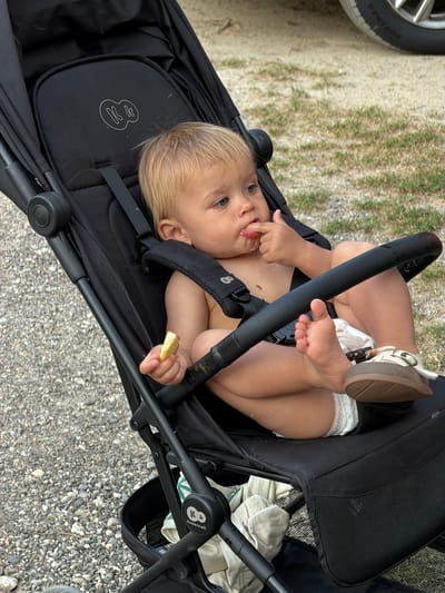 A young child is sitting in a black stroller, facing the camera. The child has blonde hair and is wearing a diaper and a small brown shoe on their right foot. They are holding a piece of food in their right hand and have their left hand near their mouth. The stroller has a black frame and black fabric, with a white logo on the headrest. The background shows a gravelly ground and some dried grass. The scene appears to be outdoors during the daytime, possibly in a park or backyard. The child's expression is one of curiosity and engagement. There is a faint logo with the word "Kinderkraft" visible on the stroller's handlebar.