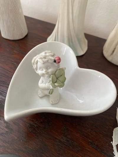 A close-up, eye-level shot shows a white ceramic heart-shaped dish containing a small, white cherub figurine. The cherub is seated and holding a green clover with four leaves. A small, red ladybug with black spots is perched on the cherub's head. The dish rests on a dark wood surface, likely a table or shelf. In the background, out of focus, are several tall, white decorative objects with ridged textures, possibly vases or sculptures. The scene is brightly lit, creating soft shadows. This image was captured in Osek, Czechia.